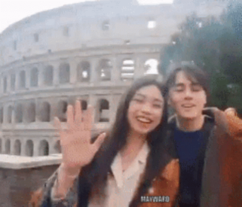 A Man And A Woman Are Waving In Front Of The Colosseum In Rome GIF