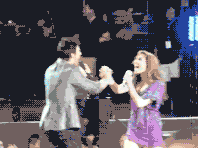 A Man And A Woman Holding Hands While Singing Into Microphones GIF