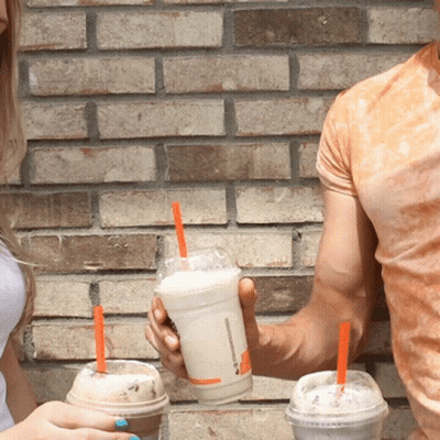 A Man And A Woman Holding Plastic Cups Of Milkshakes With Orange Straws GIF