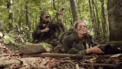 A Man And A Woman In Camouflage Are Laying In The Woods . GIF