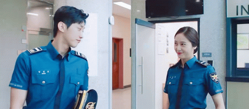 A Man And A Woman In Police Uniforms Are Standing Next To Each Other In A Building . GIF