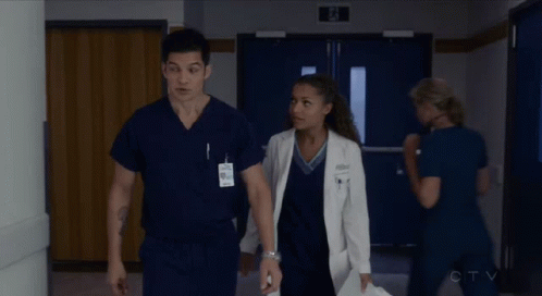 A Man And A Woman In Scrubs Are Walking Down A Hallway In A Hospital GIF