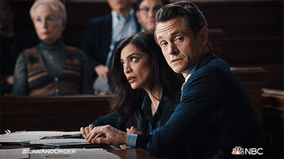 A Man And A Woman Sit At A Table In A Courtroom With #Lawandorder On The Bottom Right GIF
