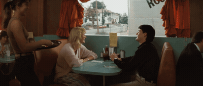 A Man And A Woman Sit At A Table In A Diner With A Sign That Says Ross GIF