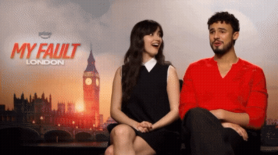 A Man And A Woman Sit Next To Each Other In Front Of A Poster For My Fault London GIF