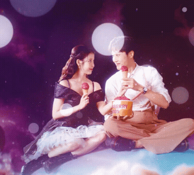 A Man And A Woman Sitting Next To Each Other With A Bucket Of Berry Ice Cream GIF