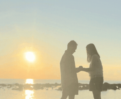 A Man And A Woman Standing On A Beach Looking At The Sunset GIF
