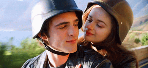 A Man And A Woman Wearing Helmets Are Hugging Each Other . The Woman Is Kissing The Man On The Cheek . GIF