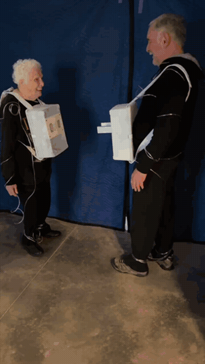 A Man And An Older Woman Are Standing Next To Each Other Wearing Cardboard Boxes On Their Arms GIF