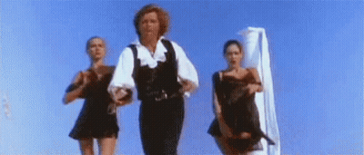 A Man And Two Women Are Dancing On A Beach In Front Of A Blue Sky . GIF
