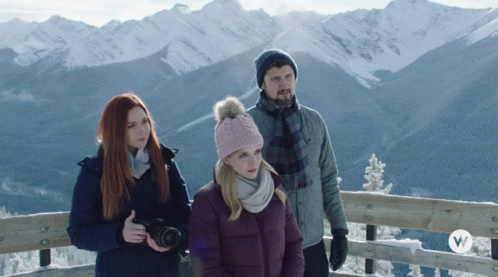 A Man And Two Women Are Standing In Front Of A Snowy Mountain Range GIF