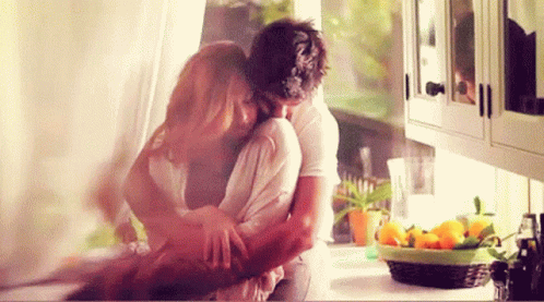 A Man And Woman Are Hugging In A Kitchen With A Basket Of Lemons In The Background . GIF