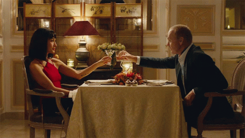 A Man And Woman Are Sitting At A Table Toasting With Wine Glasses GIF