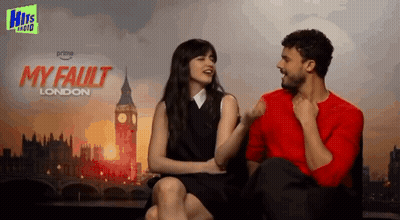 A Man And Woman Are Sitting Next To Each Other In Front Of A Poster For My Fault London GIF