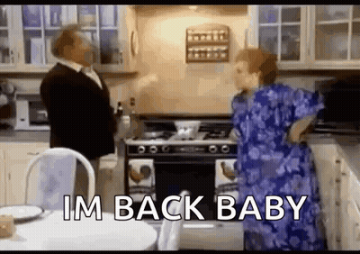 A Man And Woman Are Standing In A Kitchen With The Words `` Im Back Baby '' . GIF