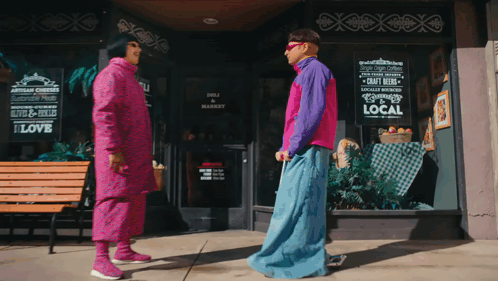A Man And Woman Are Standing In Front Of A Deli And Market Sign GIF