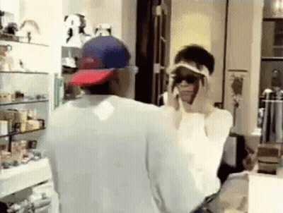 A Man And Woman Are Standing In Front Of A Mirror In A Room . The Woman Is Wearing Sunglasses And A Hat . GIF