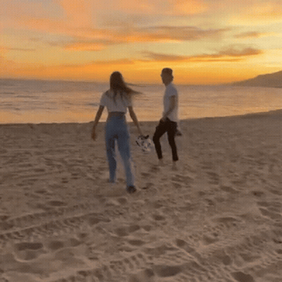 A Man And Woman Are Walking On The Beach At Sunset . GIF