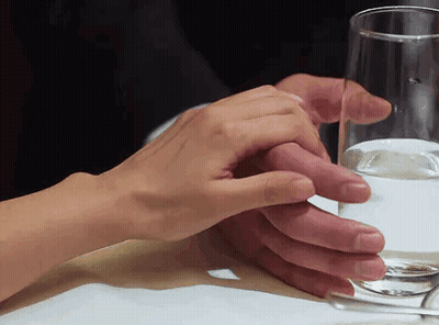 A Man And Woman Holding Hands Over A Glass Of Water On A Table GIF