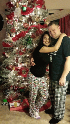A Man And Woman Pose In Front Of A Christmas Tree With The Man Wearing A Shirt That Says ' Everlast ' GIF