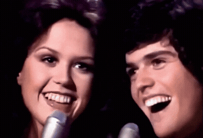 A Man And Woman Singing Into Microphones And Smiling GIF