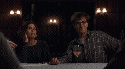 A Man And Woman Sitting At A Table With A Glass Of Wine GIF