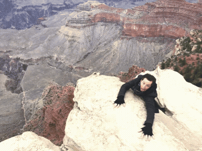 A Man Climbs Up A Rocky Cliff With A Valley In The Background GIF