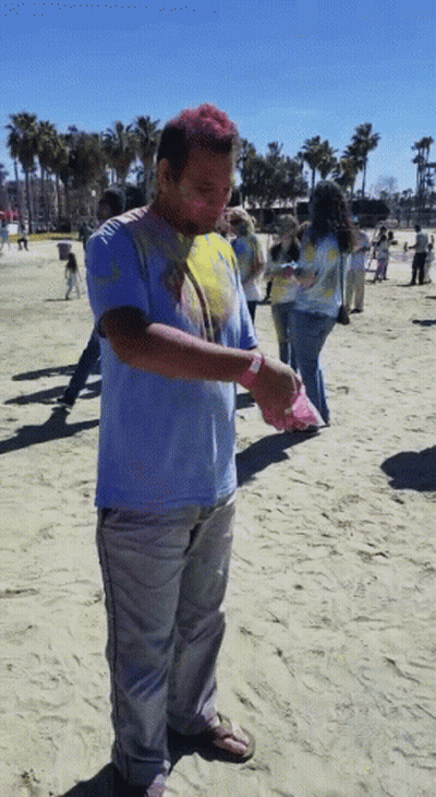 A Man Covered In Colored Powder Stands On A Sandy Beach GIF