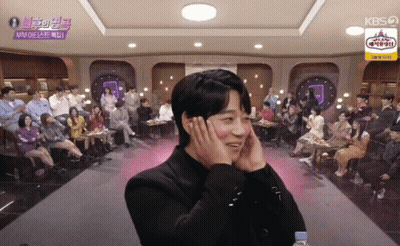 A Man Covering His Ears With His Hands In Front Of A Crowd With A Kbs Logo On The Ceiling GIF