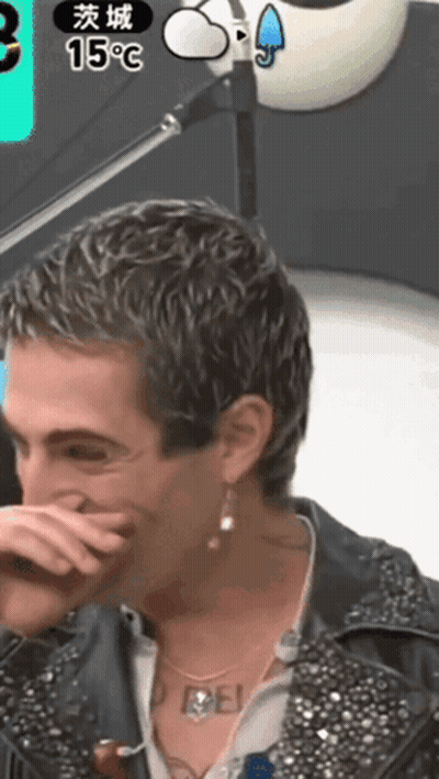 A Man Covering His Mouth With His Hand While Wearing Earrings And A Necklace That Says Rebel GIF