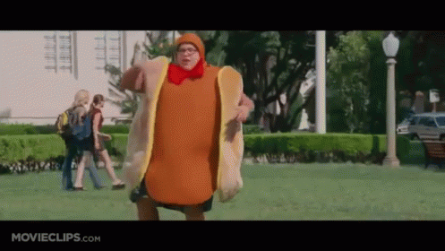 A Man Dressed As A Hot Dog Is Walking In A Park . GIF