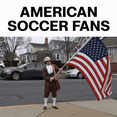 A Man Dressed As Benjamin Franklin Holds An American Flag In Front Of A Sign That Says American Soccer Fans GIF