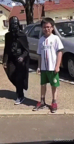 A Man Dressed As Darth Vader Stands Next To A Young Boy Wearing A White Shirt That Says Serie GIF