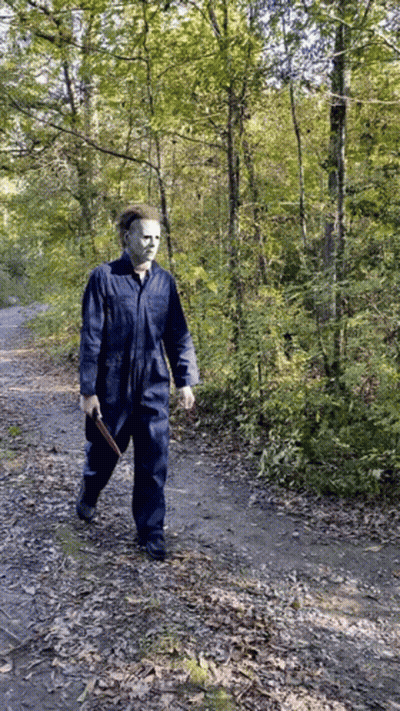 A Man Dressed As Michael Myers Is Walking Down A Path In The Woods GIF
