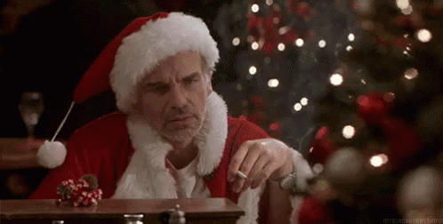 A Man Dressed As Santa Claus Is Smoking A Cigarette While Sitting At A Table In Front Of A Christmas Tree . GIF