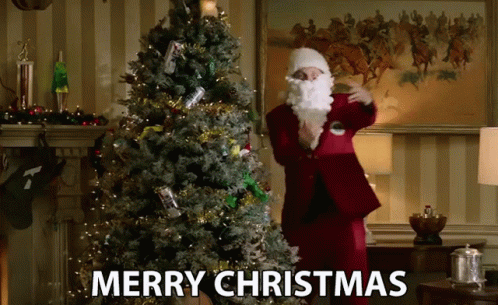 A Man Dressed As Santa Claus Is Standing In Front Of A Christmas Tree With The Words Merry Christmas Below Him GIF