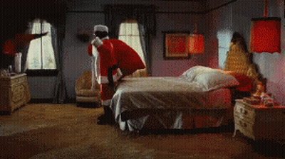 A Man Dressed As Santa Claus Is Standing On Top Of A Bed GIF