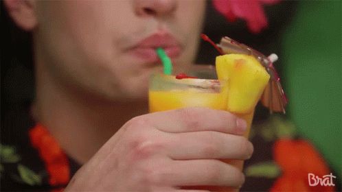 A Man Drinking A Pineapple Juice With A Green Straw From A Glass That Says Brat On The Bottom GIF