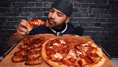 A Man Eating A Slice Of Pizza Next To A Pizza With Pepperoni On It GIF