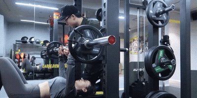 A Man Helps A Woman Lift A Barbell In A Gym With A Sign In The Background That Says Carb Dien GIF