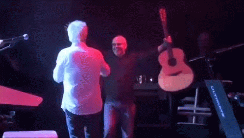 A Man Holding A Guitar On Stage With Another Man GIF