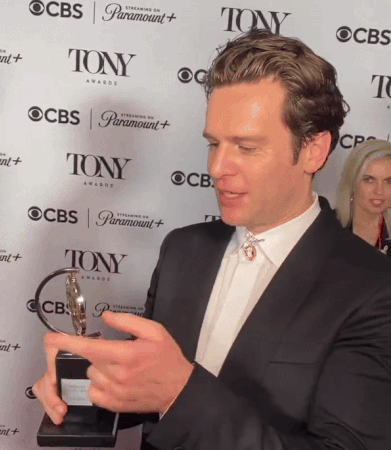 A Man Holding A Trophy In Front Of A Wall That Says Tony Awards GIF