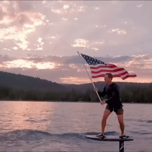 A Man Holding An American Flag While Standing On A Surfboard GIF
