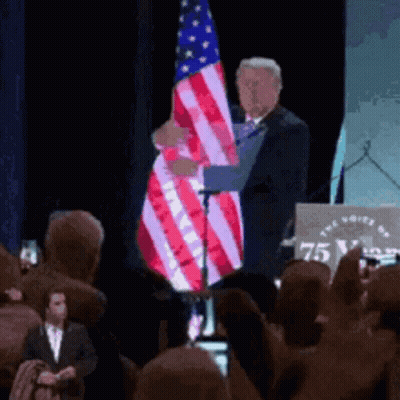 A Man Holds An American Flag In Front Of A Podium That Says 75 GIF