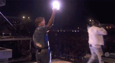 A Man Holds Up A Light While Standing On A Stage GIF