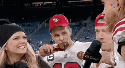 A Man In A 49Ers Hat Is Eating A Chicken Wing At A Football Game While A Woman Looks On . GIF