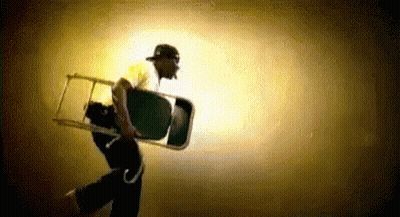 A Man In A Baseball Cap Is Carrying A Chair In A Dark Room . GIF