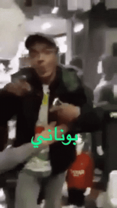 A Man In A Baseball Cap Is Holding A Bottle In Front Of A Sign That Says Botani GIF