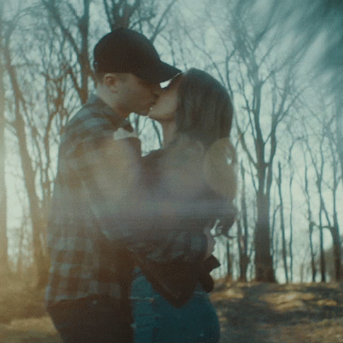 A Man In A Baseball Cap Kisses A Woman In Front Of Trees GIF