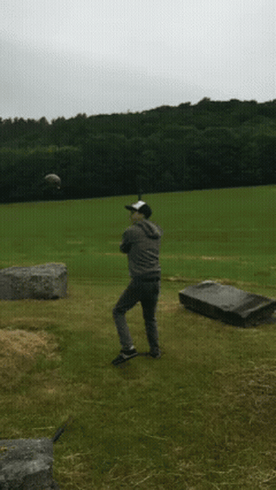A Man In A Baseball Cap Stands In A Field With A Grave In The Background GIF